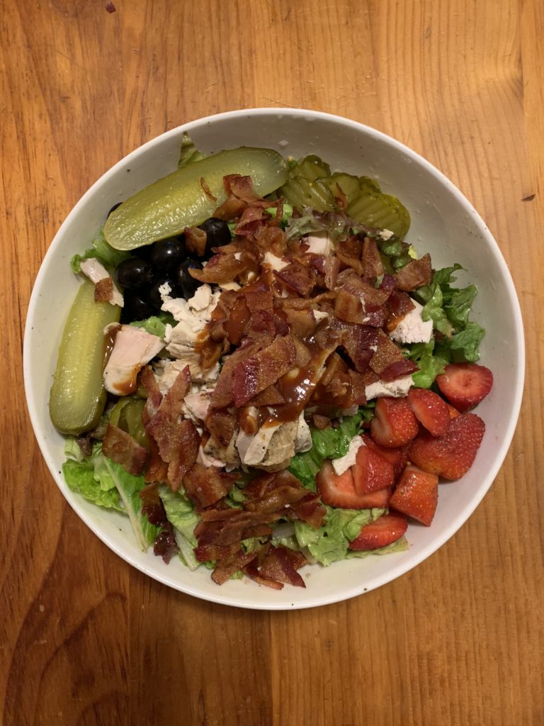 A large salad that includes farm-raised chicken, pastured bacon, real fermented pickles, fruit and olives.