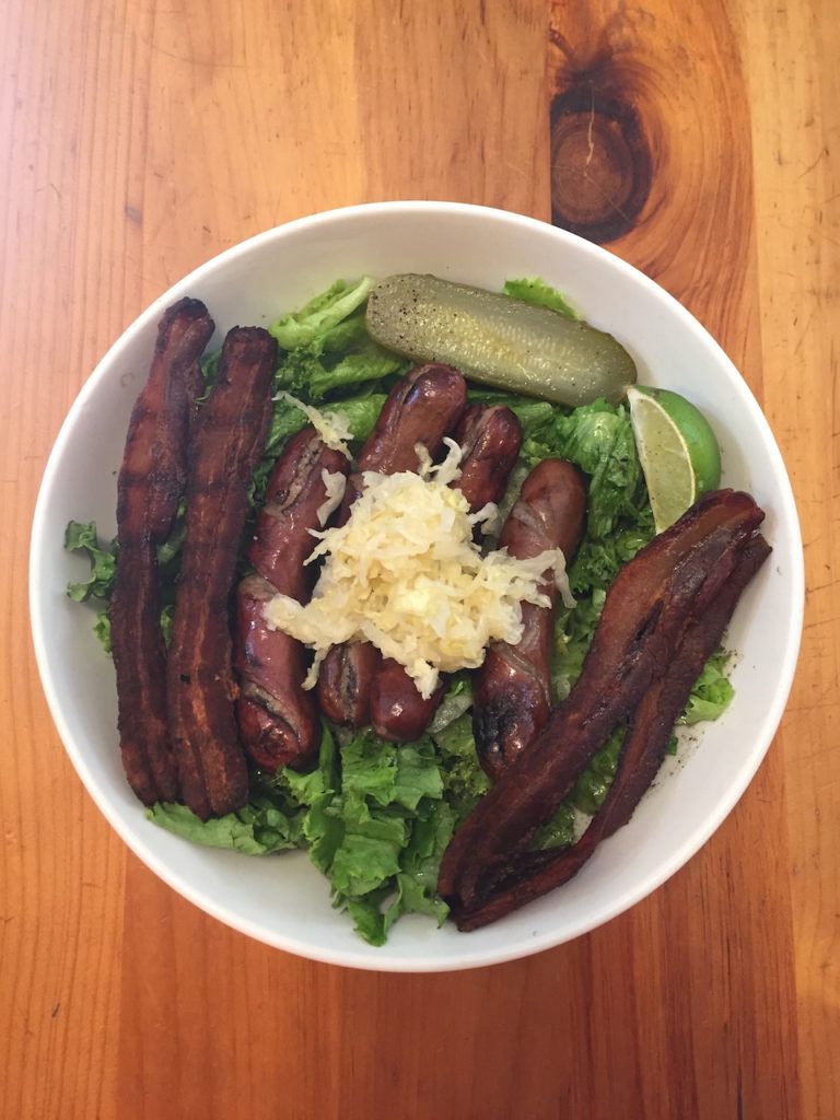 Grilled hot dogs, bacon, saurkraut and pickles over lettuce in a big bowl.