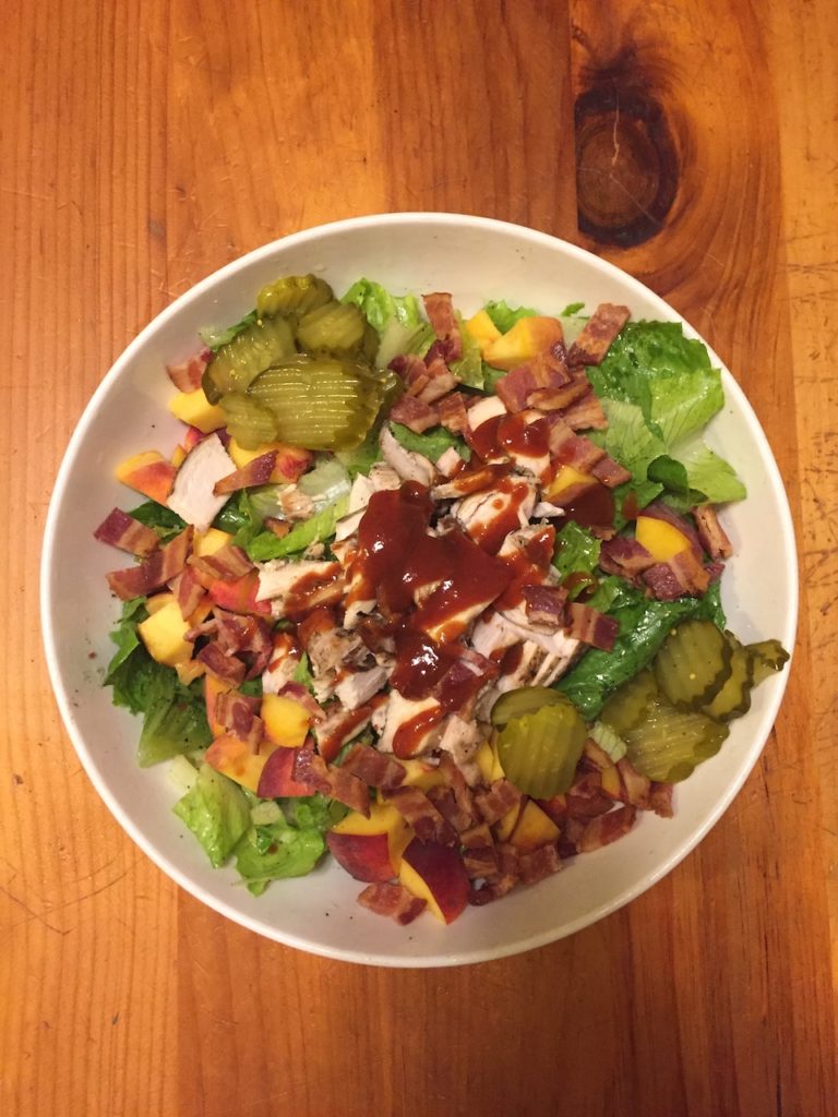 A large bowl of lettuce with chicken, bacon, fresh peaches and pickles.