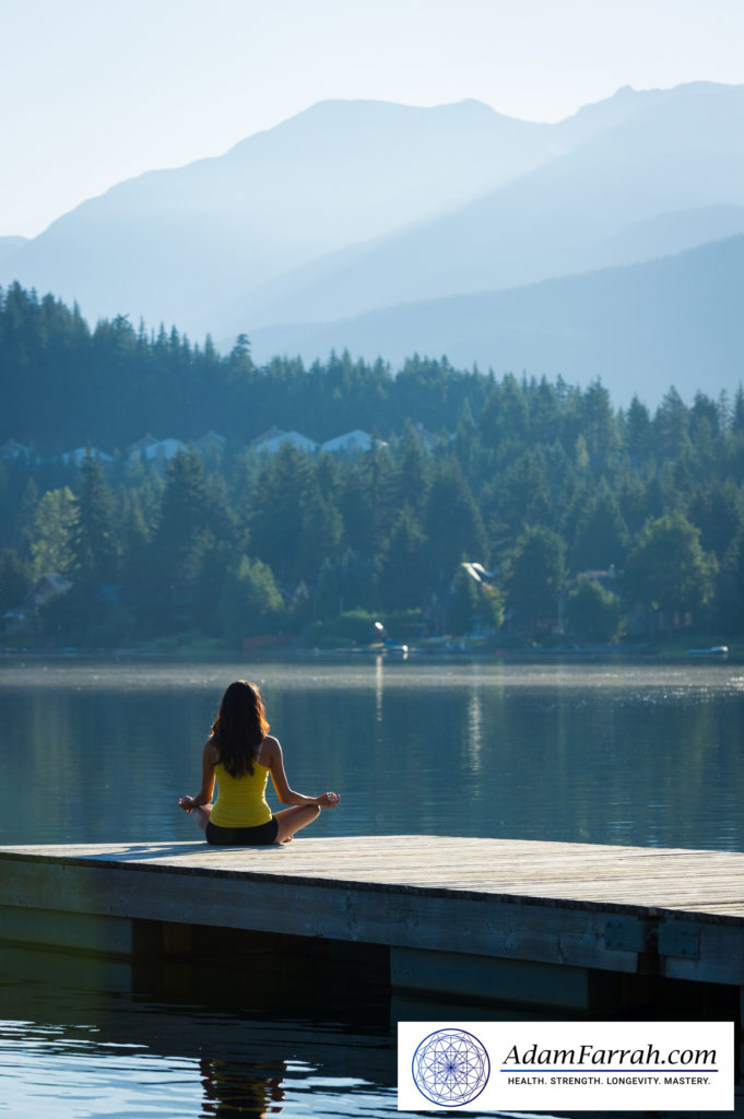 A woman seated with legs crossed meditating on a dock on a lake with trees and mountains in the background.