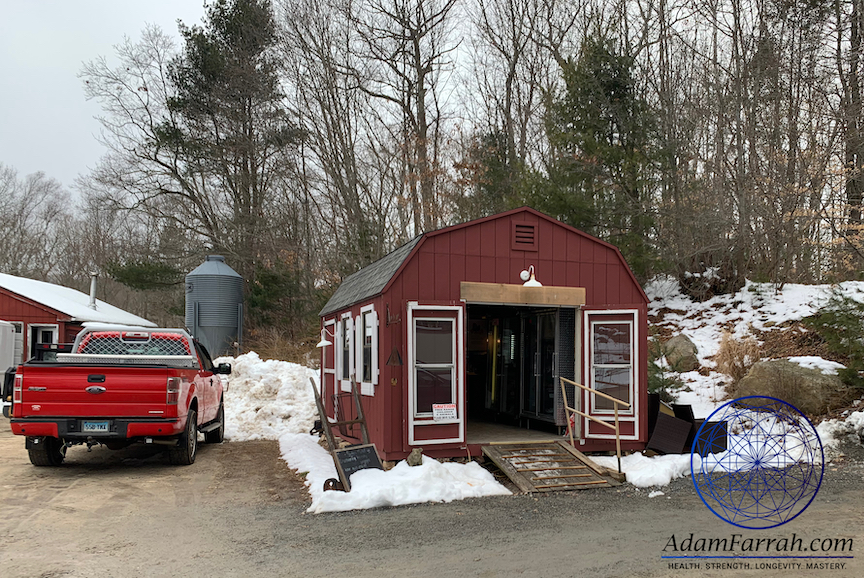 A small red farm building, a truck and a grain silo in the winter.