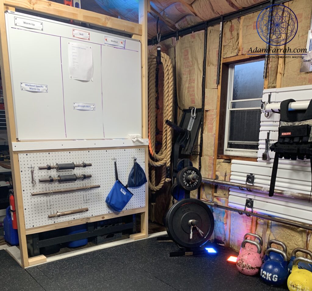A whiteboard on a wall, kettlebells, dumbbell handles, olympic plates and ropes in a garage gym.