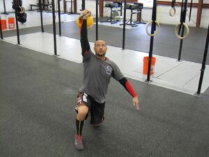 A man kneeling in a gym with a Kettlebell pressed overhead in the kneeling portion of the Kettlebell Turkish Get-Up.