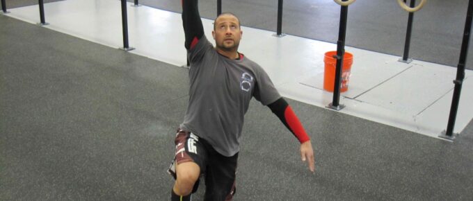 A man kneeling in a gym with a Kettlebell pressed overhead in the kneeling portion of the Kettlebell Turkish Get-Up.